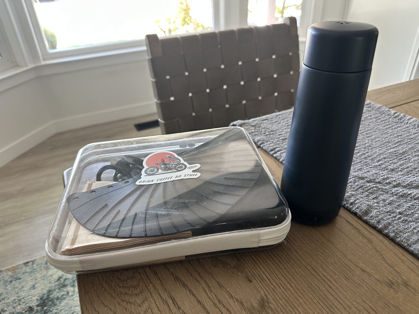 The travel coffee kit: a flat pour-over dripper and hand grinder zipped into a clear case, next to a black cylindrical electric kettle, on a wooden table by a window.