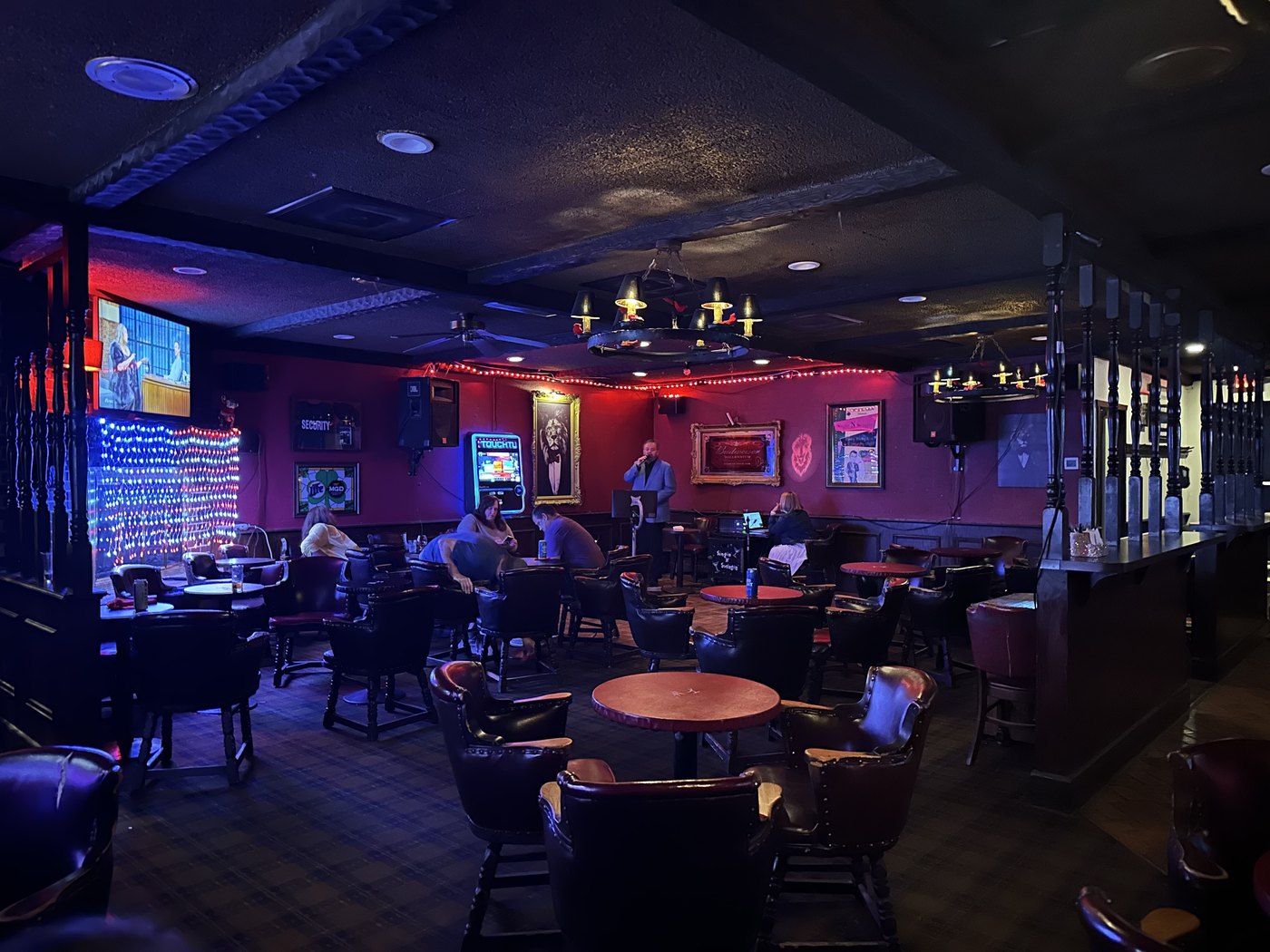 The Red Lion’s interior: red walls under low light, a string of red bulbs, a framed lion painting, and someone at the mic in front of the karaoke setup.